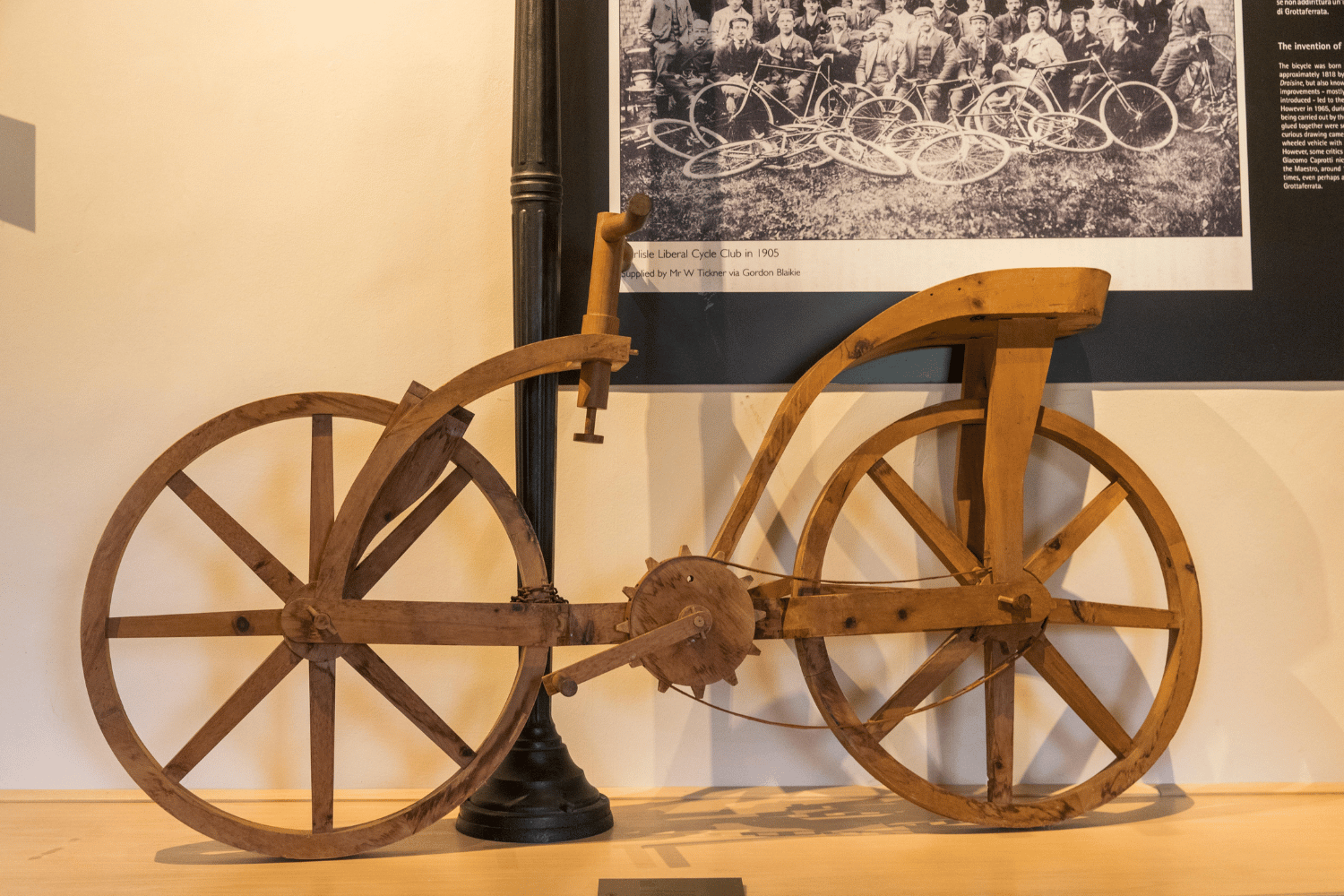 Bicicletta lignea con manubrio sospeso connesso a ruota anteriore, sella inclinata fino ai pedali, corda al posto della catena.